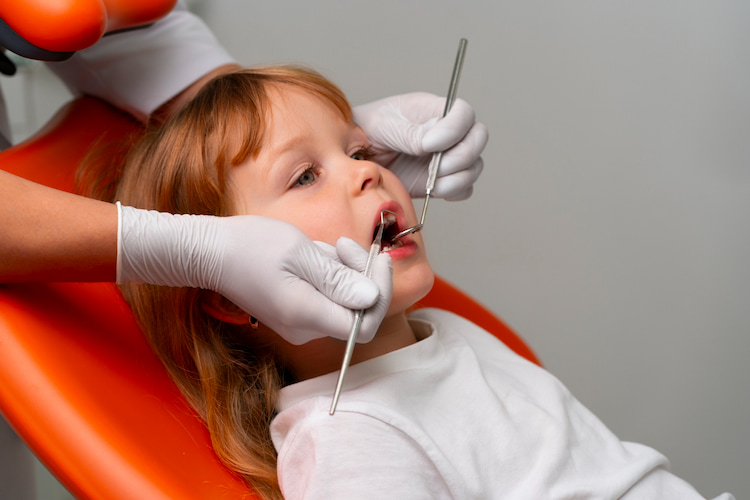 a child is sitting on a dental chair calmly and a dentist is checking her teeth using dentistry tools.