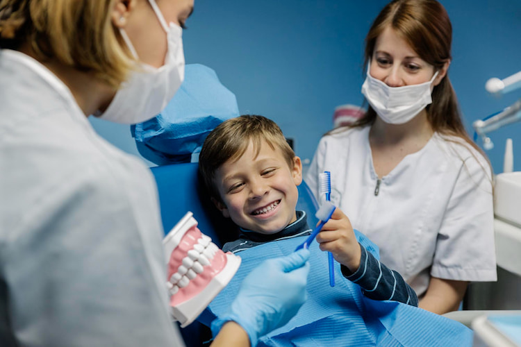 A smiling young boy in a dental chair holds a toothbrush while two masked dental professionals demonstrate on a model mouth.